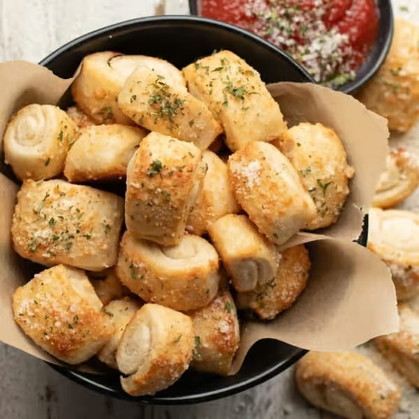A black bowl filled with golden-brown garlic knots, garnished with herbs and parmesan cheese, with marinara sauce in the background.
