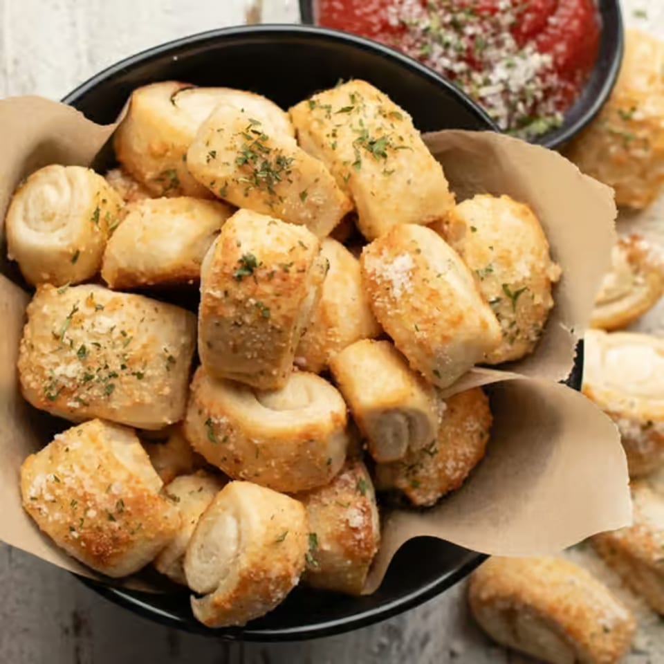 A black bowl filled with golden-brown garlic knots, garnished with herbs and parmesan cheese, with marinara sauce in the background.